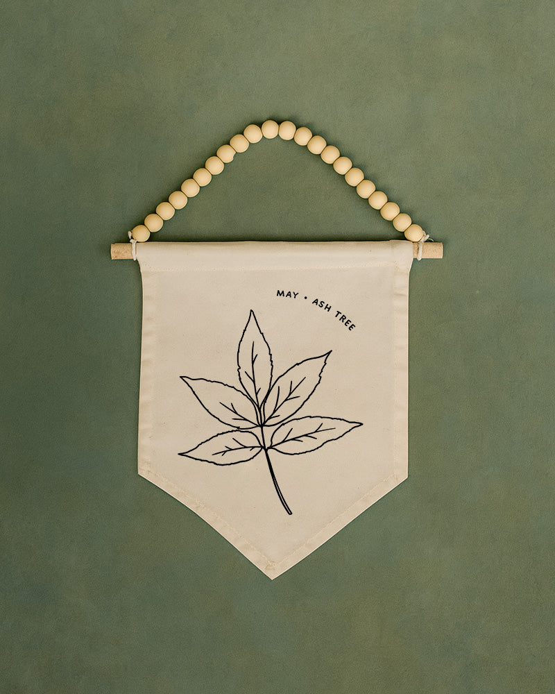 A beaded banner with a leaf design and the text "MAY - ASH TREE" printed on it.