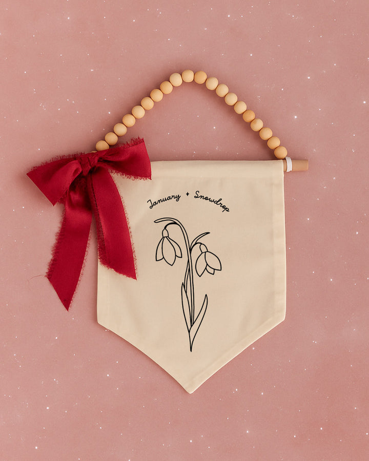 A banner with a floral design hangs against a pink background, accompanied by a red ribbon and wooden beads.