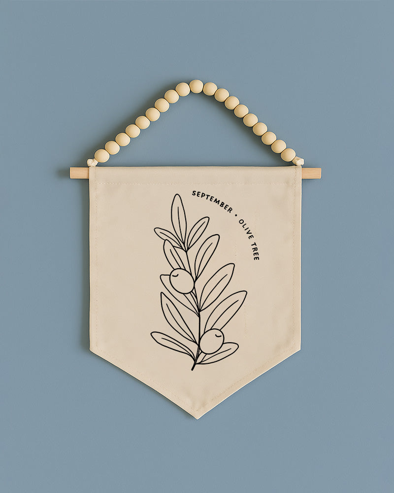 A beige banner with a black line drawing of an olive tree and the text "September olive tree" hanging on a wooden bead.