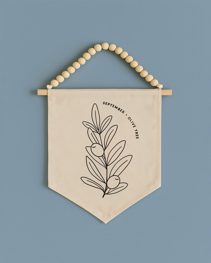 A beige banner with a black line drawing of an olive tree and the text "September olive tree" hanging on a wooden bead.