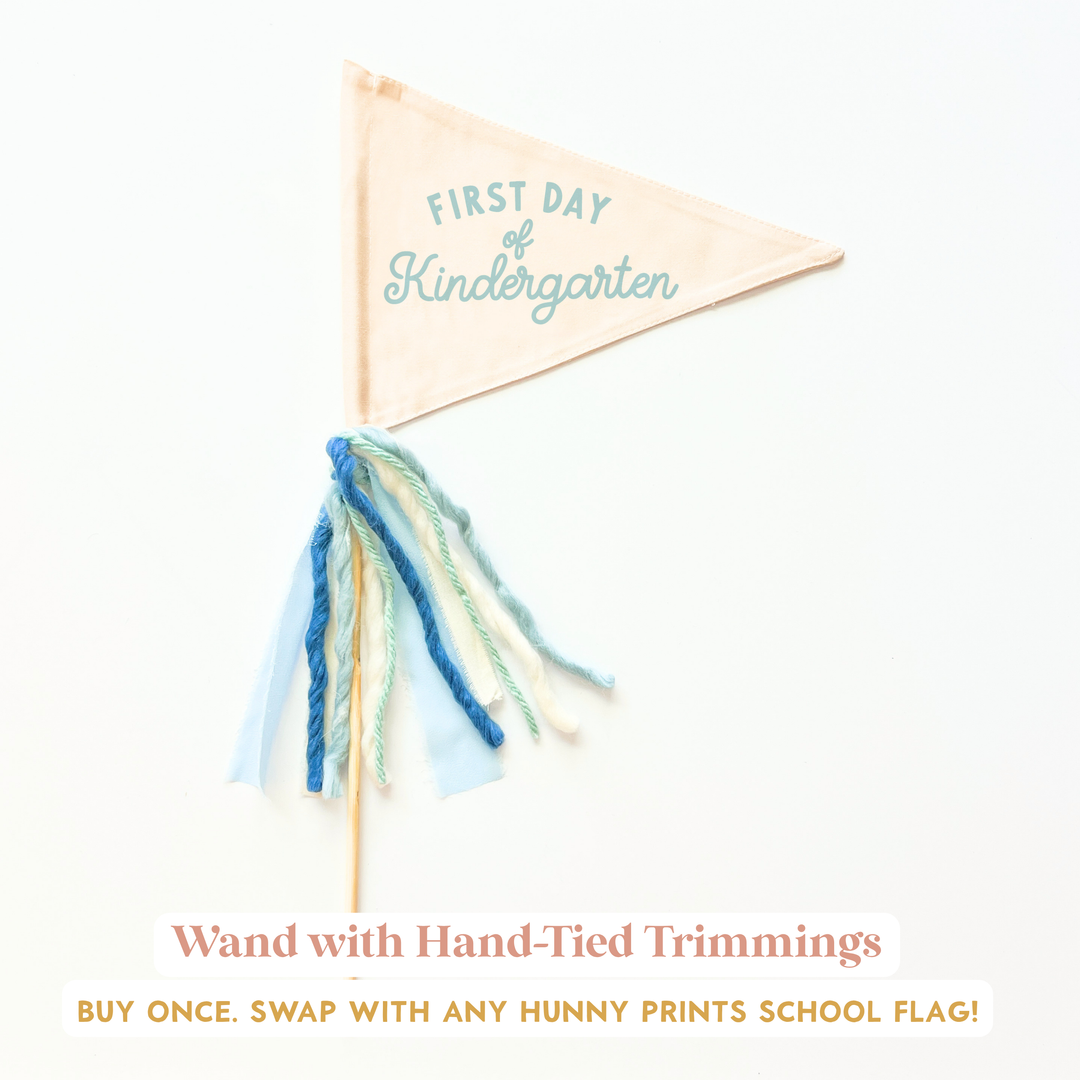 a small, beige-colored triangular flag with the text "FIRST DAY of Kindergarten" printed on it. The flag has a blue and white tassel attached to it.