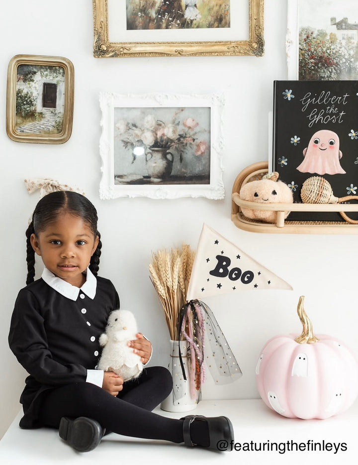 Young girl in a black outfit holding a white plush toy, sitting on a white surface with Halloween decorations in the background.
